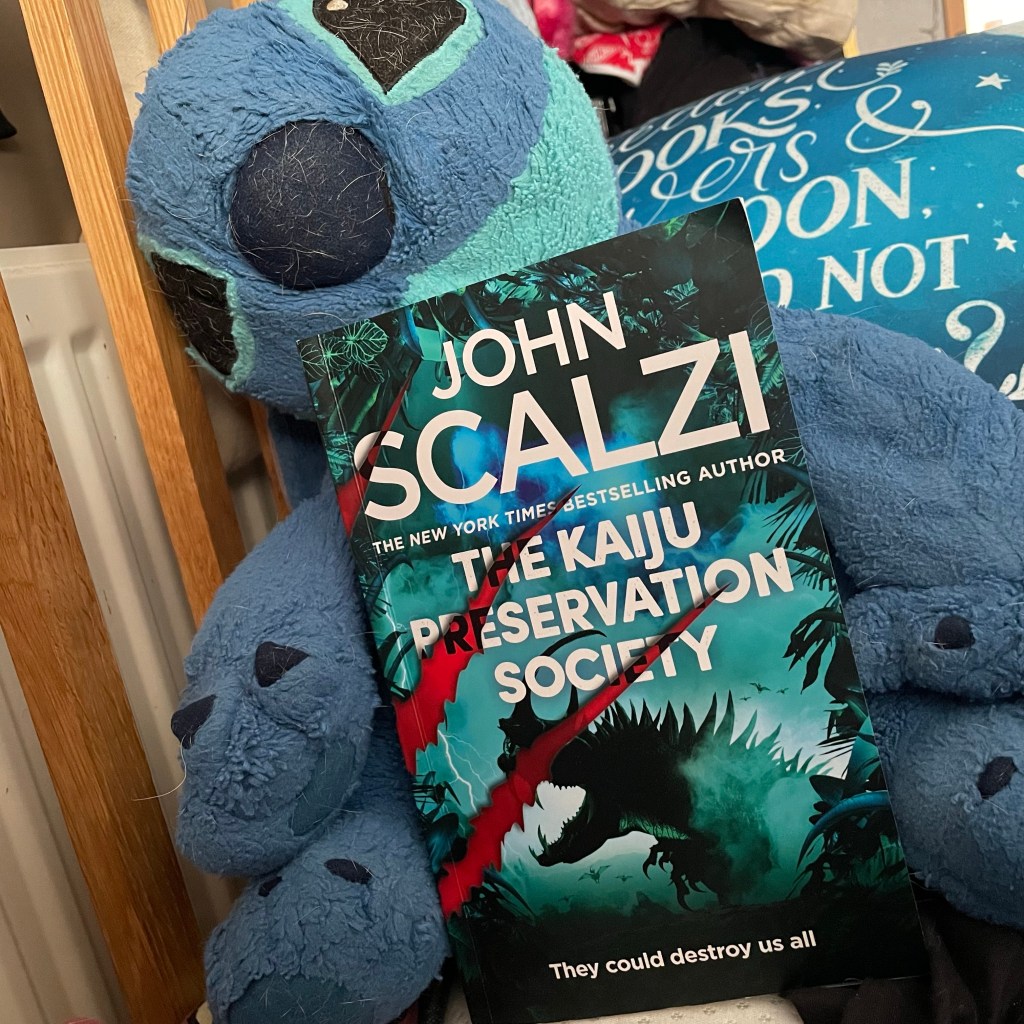 A picture of a plushie character of Disney’s Stitch (covered in dog hair) holding a blue Advanced Reading copy of the book The Kaiju Preservation Society by John Scalzi. The book cover has three red slashes - like claw marks- over a picture of a Godzilla like creature. 